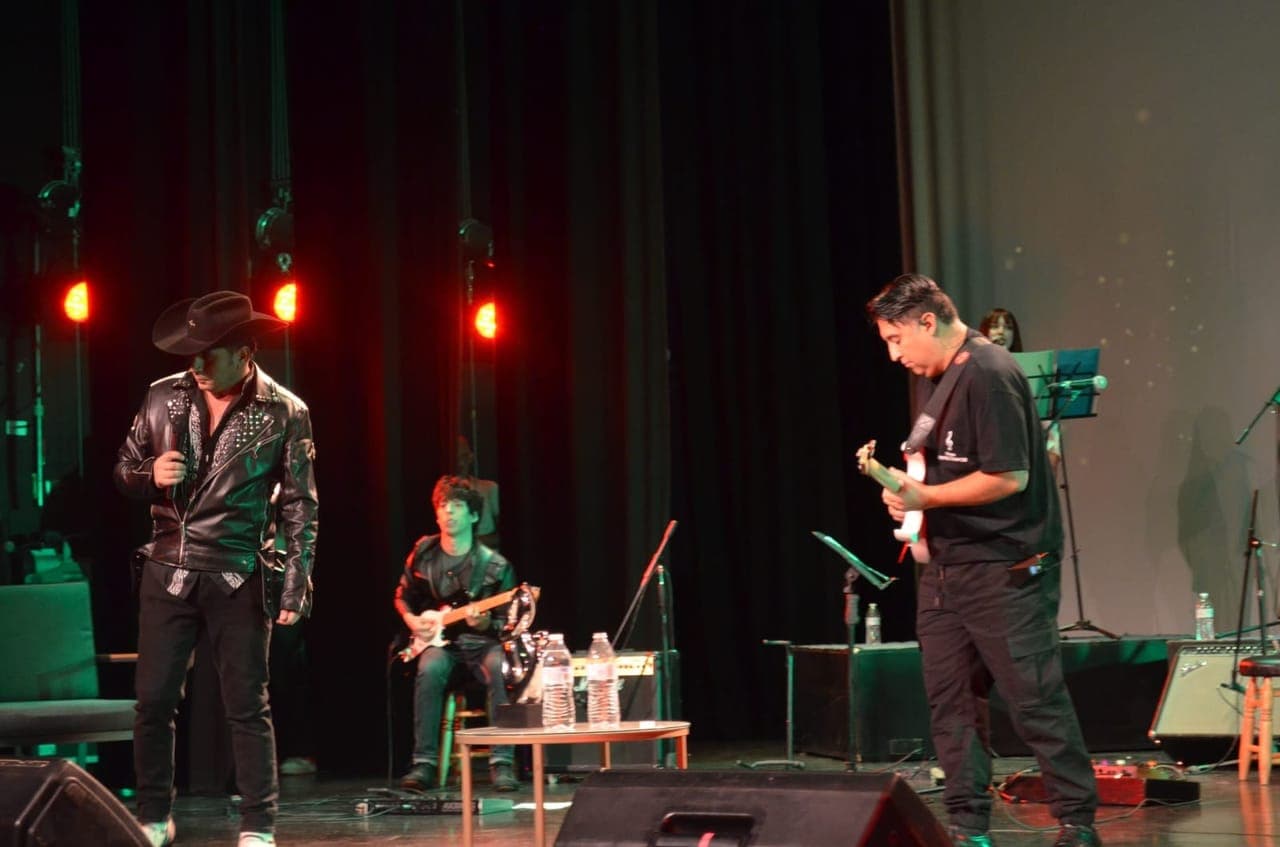 Show en teatro con luces rojas, cantante con sombrero y guitarristas